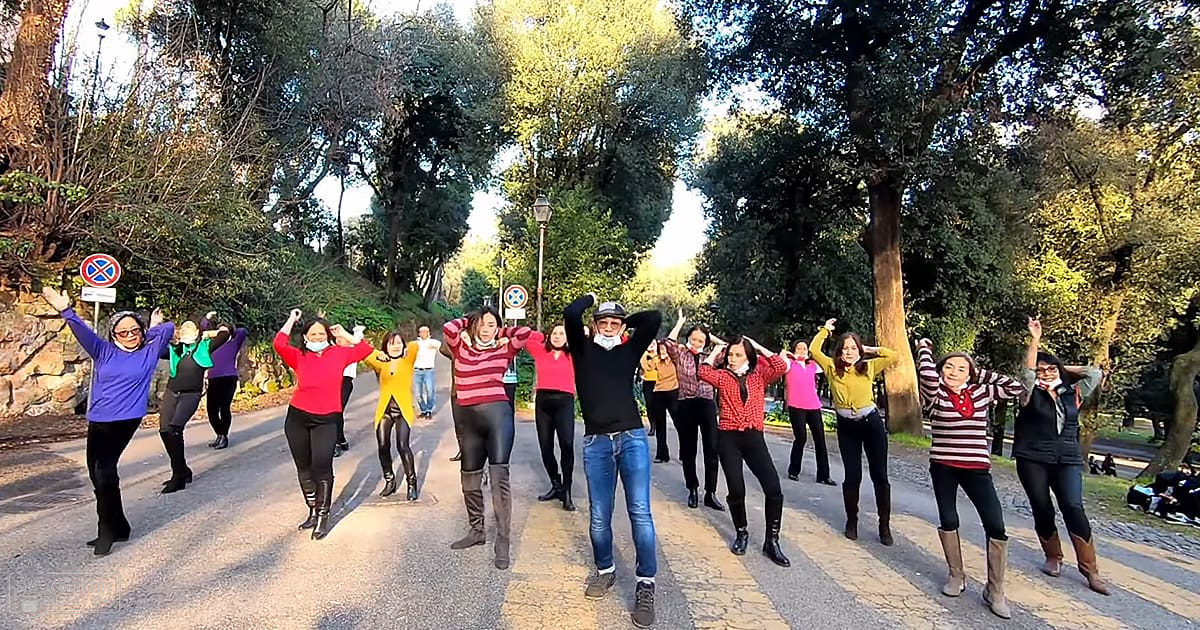 Adult females in various casual outfits participating in an outdoor activity under trees with pathways, suggesting a park setting.