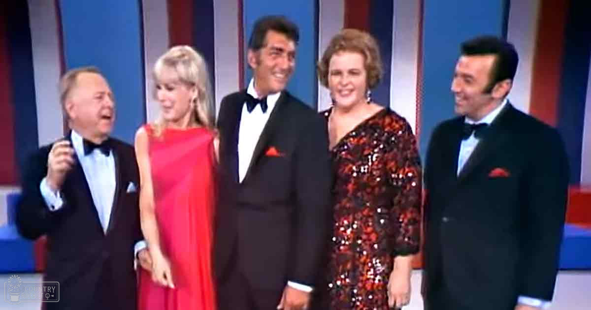 A group photo taken indoors at what appears to be a television show or formal gathering; adults in tuxedos and dresses with red accents, set against vertical striped backdrop of blue, red, and white.