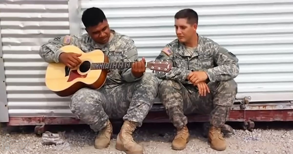 Two young adult male soldiers playing music in an outdoor setting with a corrugated metal wall and gravel or dirt ground.