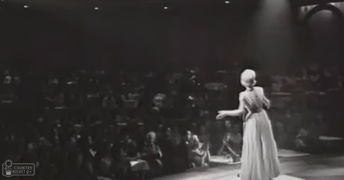 An adult female performer in a vintage-style long dress with a fitted bodice and flared skirt, performing on stage under focused lighting.