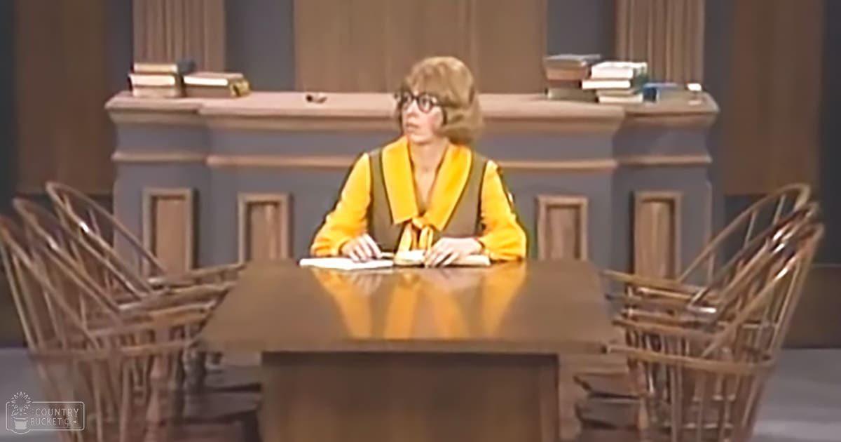 A middle-aged woman wearing a yellow high-necked blouse with long sleeves and a dark vest over it, sitting at a wooden table in what appears to be an indoor courtroom setting.