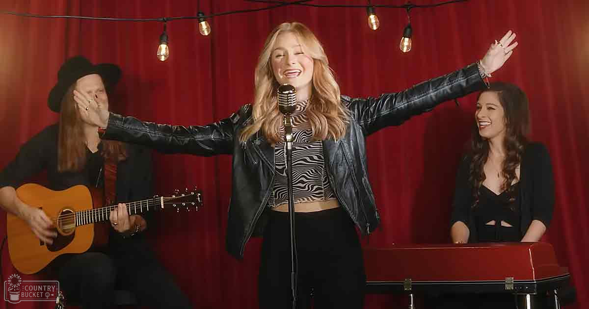 Young adult female singing into a microphone, wearing a black jacket and hat in an indoor music performance setting with string lights and red curtain backdrop.
