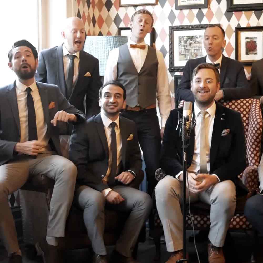 Group of men in formal attire at an indoor event with patterned chairs and framed pictures on the wall.