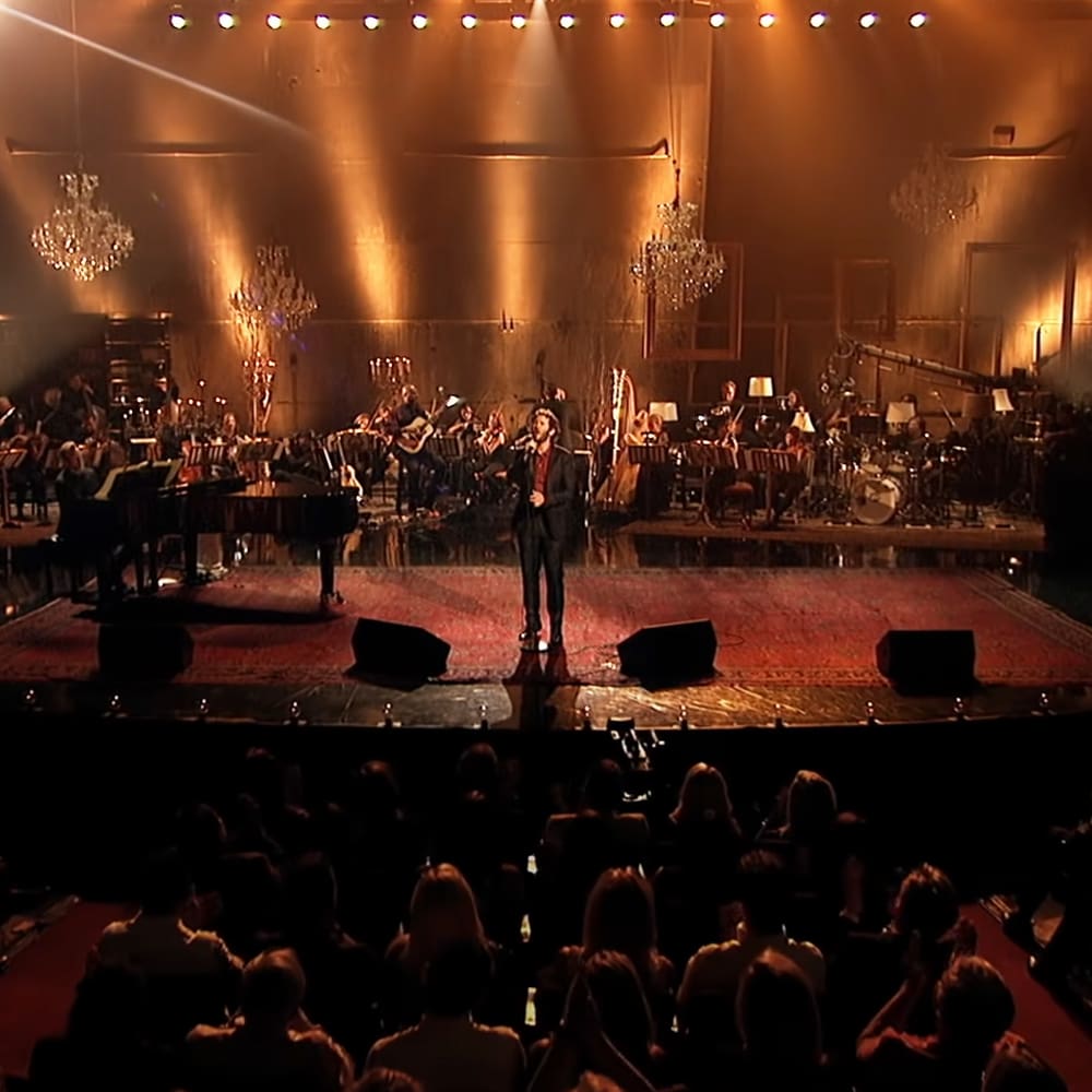 Adult male singer performing on center stage at a concert hall with an orchestra behind him; evening artificial lighting, warm tones of gold, brown, red, black attire.