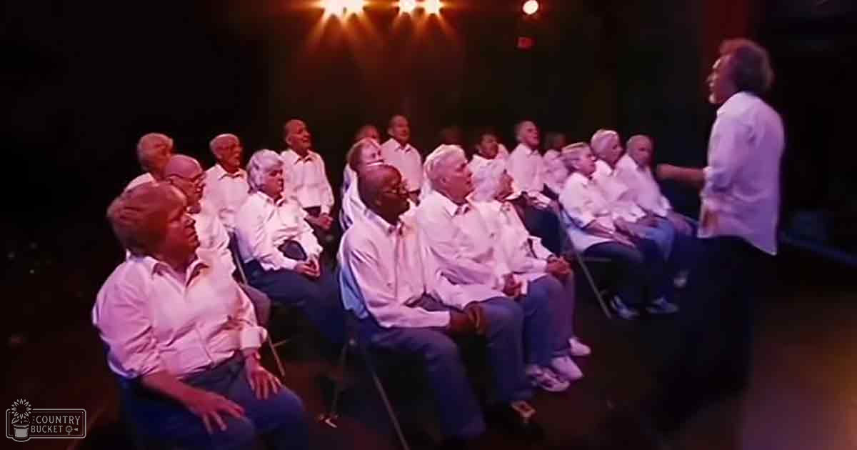 A group of older adults in white button-up shirts, seated in rows in a dimly lit indoor space resembling a theater or performance venue.