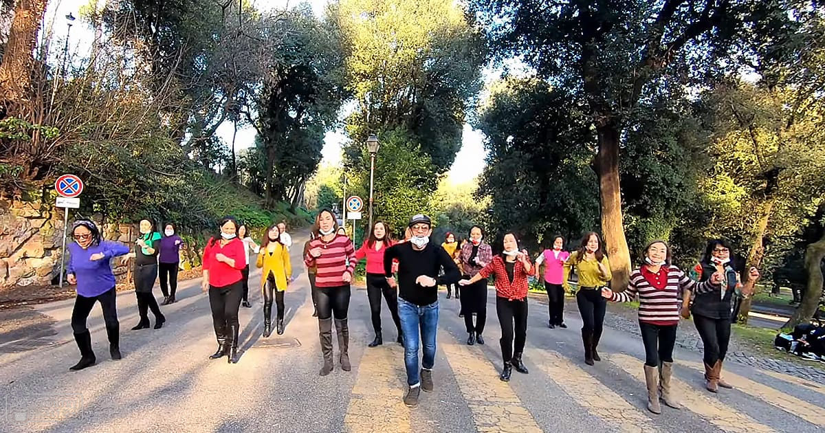 Adult females dancing outdoors on a paved path surrounded by greenery in what appears to be a park or wooded area. The scene is bathed in the soft light of late afternoon or early evening, with shadows indicating the angle of sunlight.