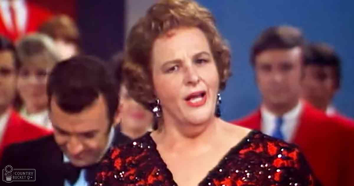 Adult woman with short, styled hair in a red sequined dress at an indoor event. Background includes formally dressed individuals and a blue backdrop suggesting a stage or studio setting.