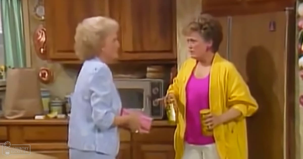 Two middle-aged to older adult women in a kitchen setting, wearing light blue button-up shirts and bright yellow cardigans over pink tank tops. One holds a yellow cup and what appears to be a small pink box or package.