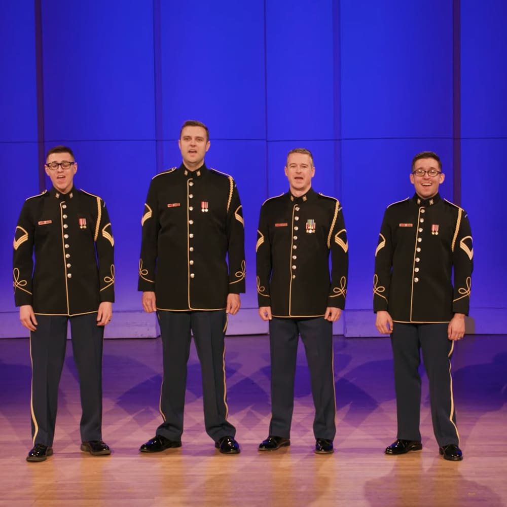 Young adult male singers in uniform military-style jackets with red ribbons, performing on a wooden stage under blue and purple lighting.