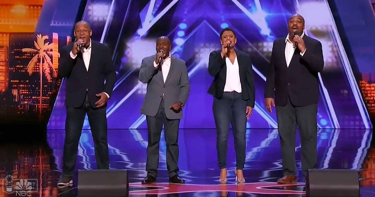A talent show or competition performance on a stage with a singing group in suits against a vibrant cityscape backdrop with palm trees and illuminated buildings, under dynamic lighting at night.