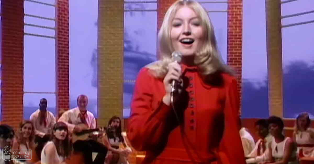 A young adult female singer wearing a red vintage-style dress performs into a microphone, accompanied by a band of young adults and middle-aged individuals in a well-lit studio with brick columns and outdoor scenery visible through the windows.