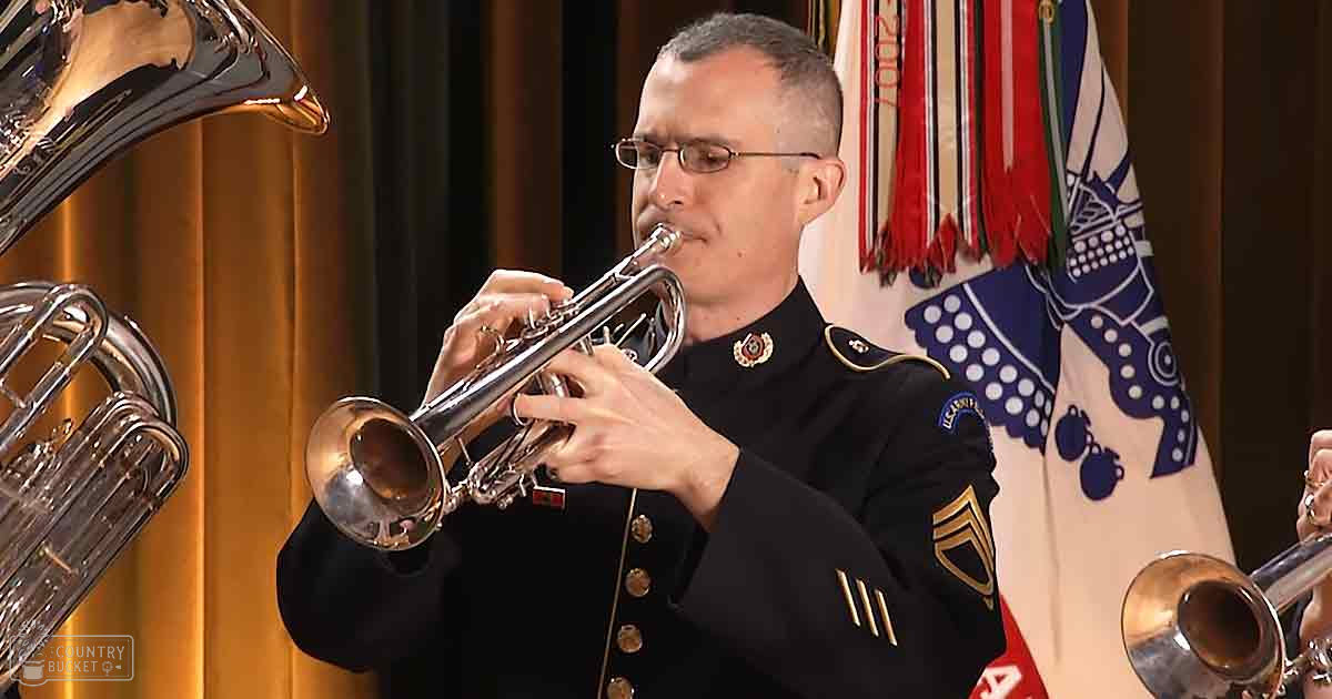 A middle-aged male wearing a dark military uniform with gold insignia, playing a trumpet indoors during what appears to be a formal event with colorful flags hanging in the background.