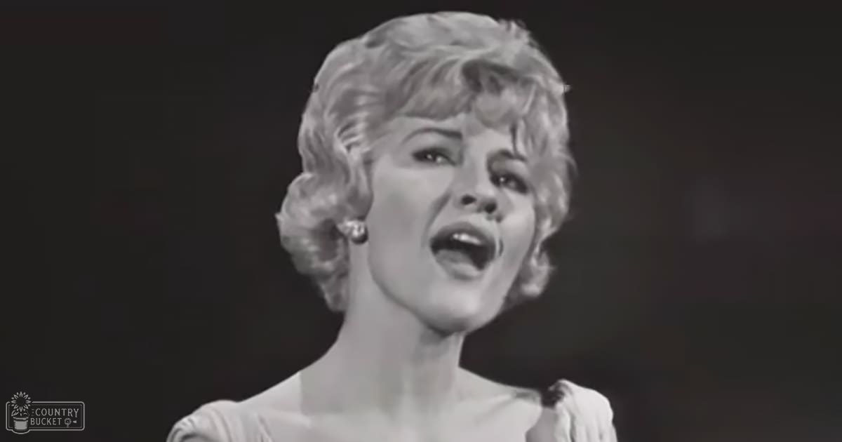 A middle-aged woman singing in a performance setting with a dark stage backdrop and monochromatic lighting.