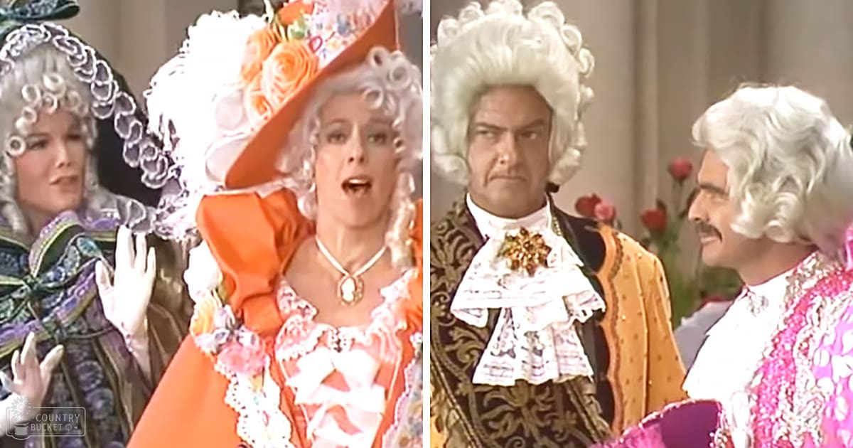 Historical drama or opera performance set in 18th-century Europe, with a vibrant orange dress and black and gold brocade jacket adorned with intricate patterns and floral decorations.
