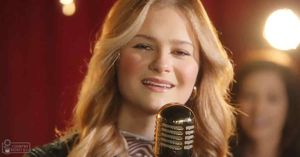A young adult female sings into a vintage-style microphone, wearing a striped dress with low-cut neckline and short sleeves. The setting suggests an indoor stage or performance area with red curtains.