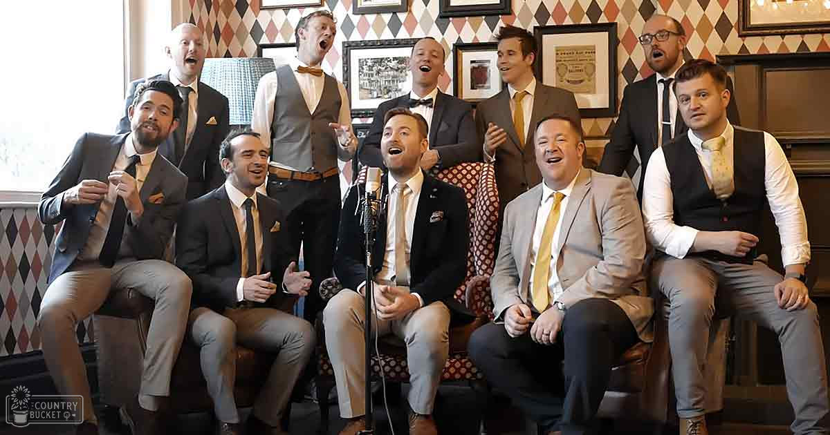 A group of men singing at an indoor event with patterned wallpaper and framed pictures in the background.