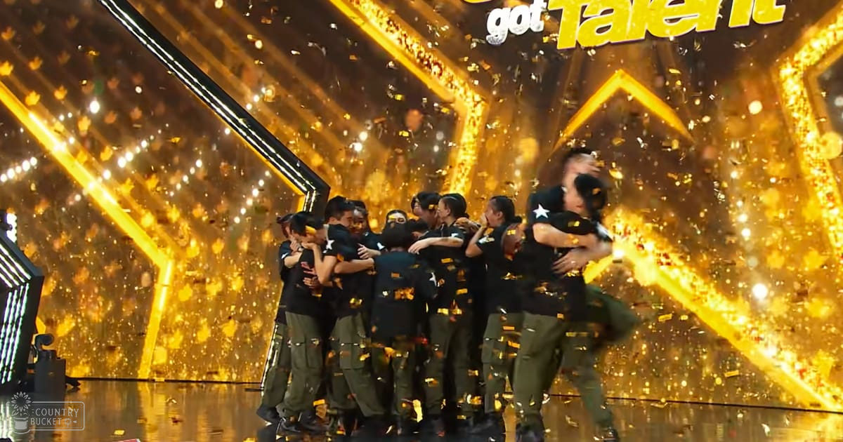 A group of young adults to middle-aged adults celebrate a win at 'Got Talent' with confetti falling around them, dressed in matching black t-shirts and olive green cargo pants on a brightly lit stage.