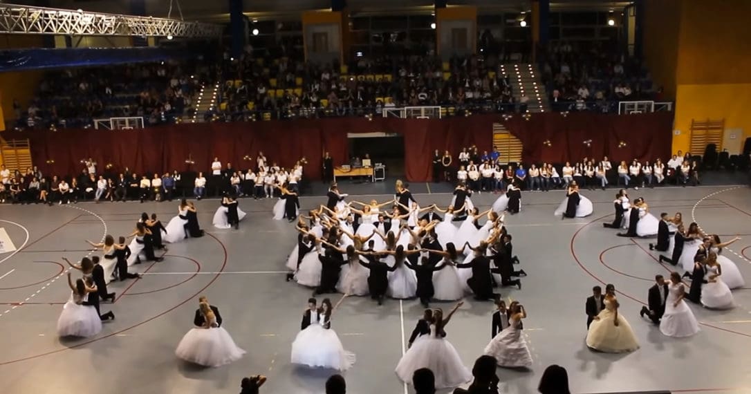 Young adults perform a dance routine indoors, surrounded by an audience seated on bleachers under artificial evening lighting. The setting is vibrant with white dresses and black tuxedos contrasting against the gray indoor arena floor.