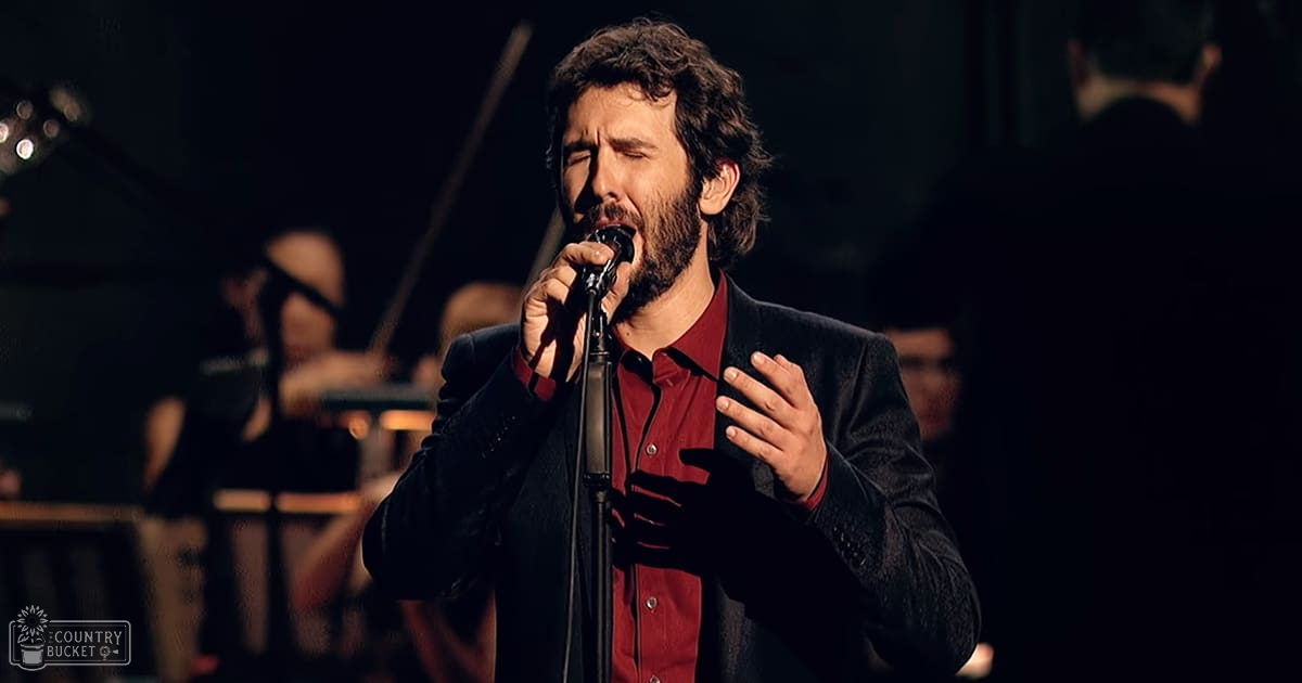 A middle-aged male singer performing on stage with a microphone stand and an orchestra in the background, dimly lit during evening or night.