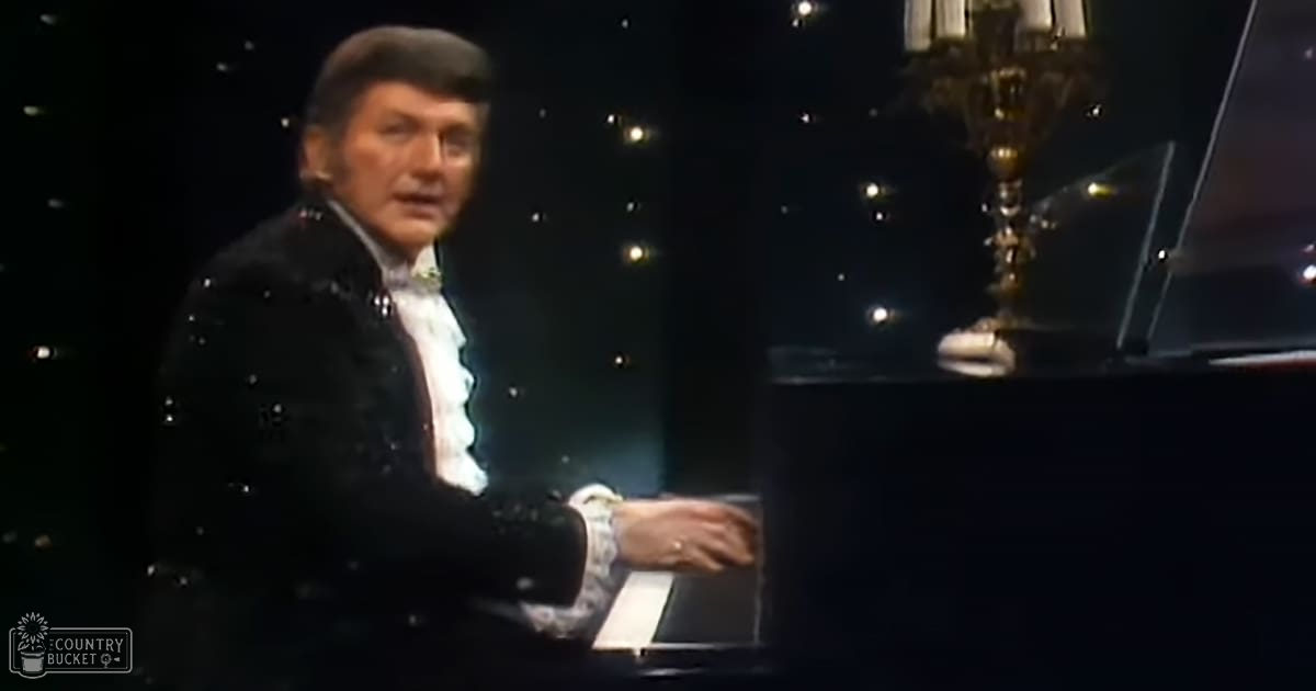 A middle-aged male performer wearing a black sequined jacket, white ruffled shirt with cufflinks, on stage in an indoor concert hall setting.