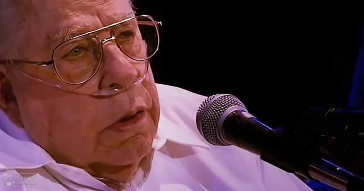 An older adult male performer wearing a white collared shirt, speaking into a microphone on an indoor stage with focused lighting.
