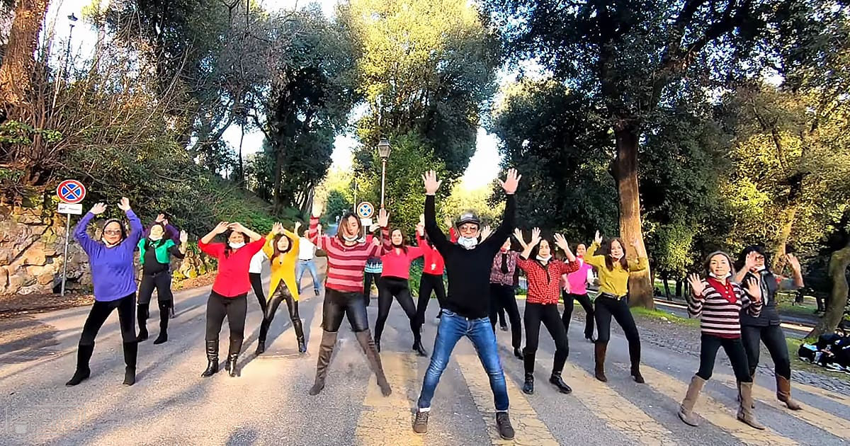 A group exercise class outdoors in a park-like setting with trees, paved path, and road signs indicating no entry; spring or summer foliage suggests daytime sunlight.