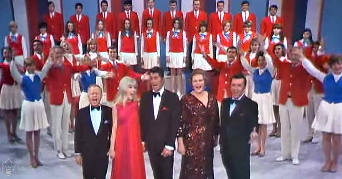 A group performance scene indoors with adults in formal attire and children in matching red jackets. The setting appears to be a stage with no visible furniture or home decor.