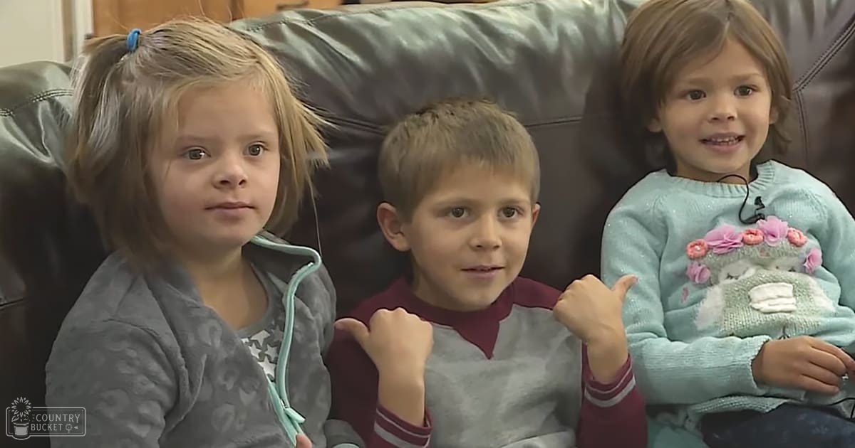 A group of children sitting together indoors, including a toddler girl in a gray fleece jacket with a light blue zipper and patterned inner layer, a child boy wearing a maroon and gray long-sleeve shirt, and another toddler girl in a light blue knitted sweater holding a pink and white stuffed animal. The setting appears to be a living room with a dark gray leather sofa.