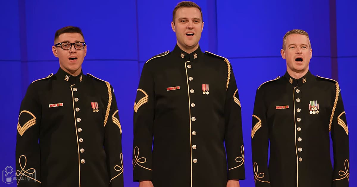 Three men in military uniforms performing indoors under stage lighting, with a blue backdrop suggesting an event or show.
