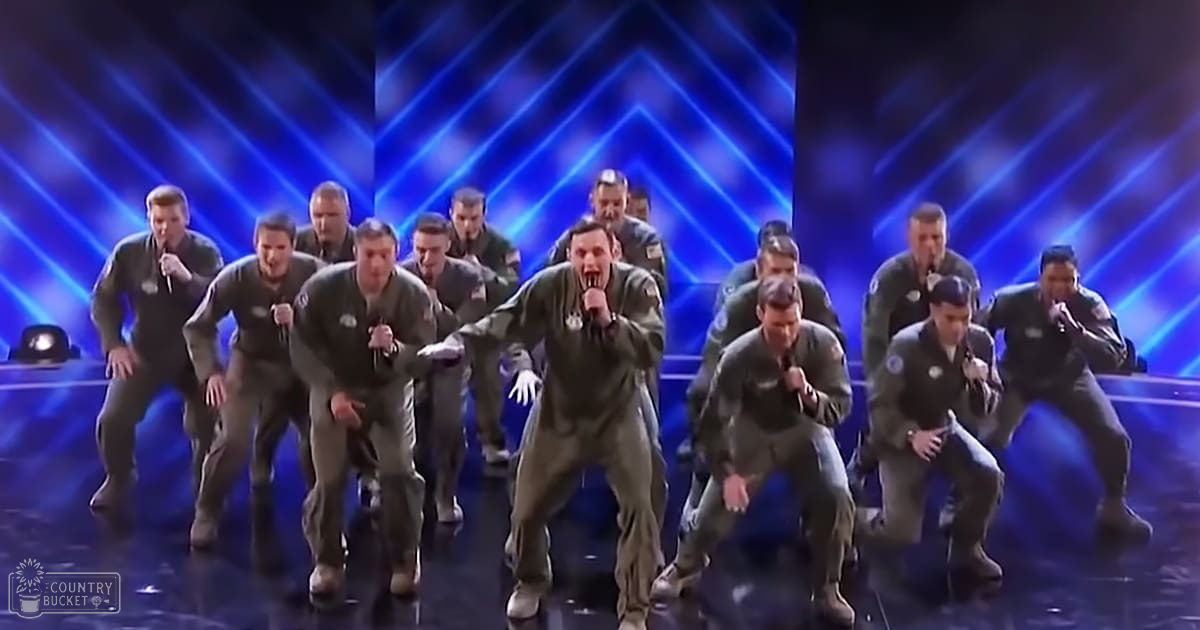 A group of young adult male performers in a stage setting, wearing matching military-style flight suits with patches, performing under blue stage lights.