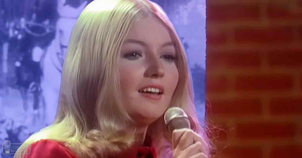 A young adult female sings into a microphone against a brick wall and a blurred poster of a horse-drawn carriage, suggesting an indoor performance venue with controlled lighting.