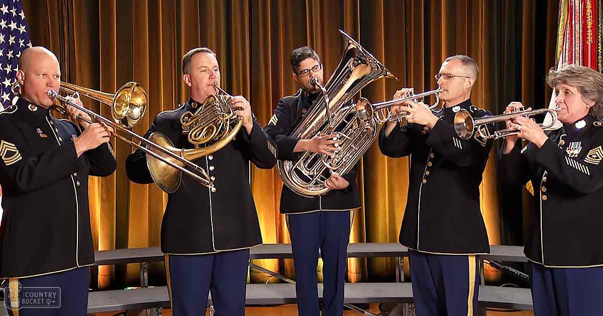 A group of adult male musicians in dark military-style jackets perform on an indoor stage with golden curtains and flags, under artificial light.