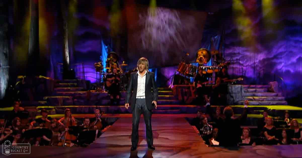 A male singer performing on a stage in an indoor concert hall, surrounded by musicians and stage lighting.