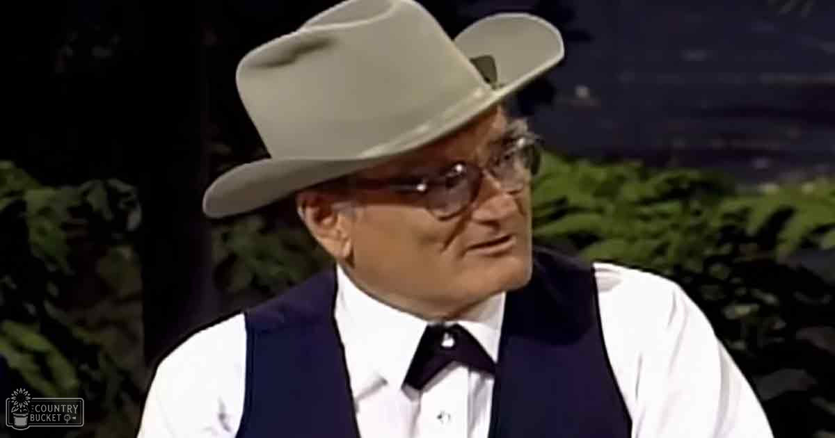 An older adult male wearing a white cowboy hat and glasses is seated in what appears to be a studio setting with controlled lighting. The environment suggests an indoor setup, possibly for a talk show or interview.