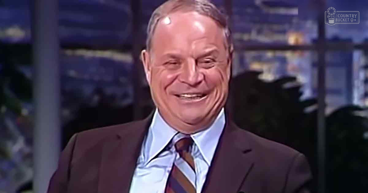An older adult male wearing a dark suit jacket over a light blue dress shirt with a striped tie in a studio setting.