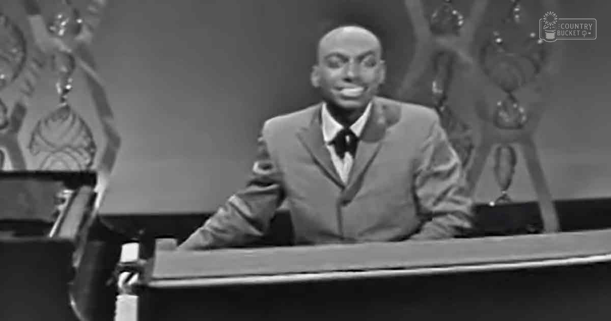 Adult male in a suit at an indoor studio or performance setting with a grand piano and stage-like background elements.