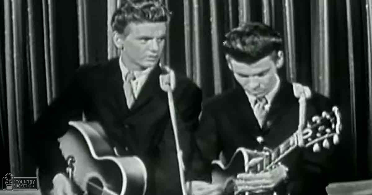 Two young adult male musicians in formal suits from the mid-20th century performing or recording music on a stage with a microphone and curtain backdrop.