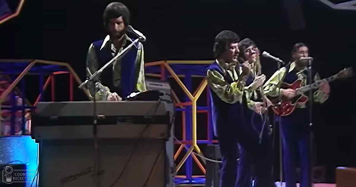 A musical performance scene with four adult male performers, each wearing dark-colored outfits with light-colored patterned shirts, set against an indoor venue with stage lighting and a keyboard or synthesizer visible on the stage.