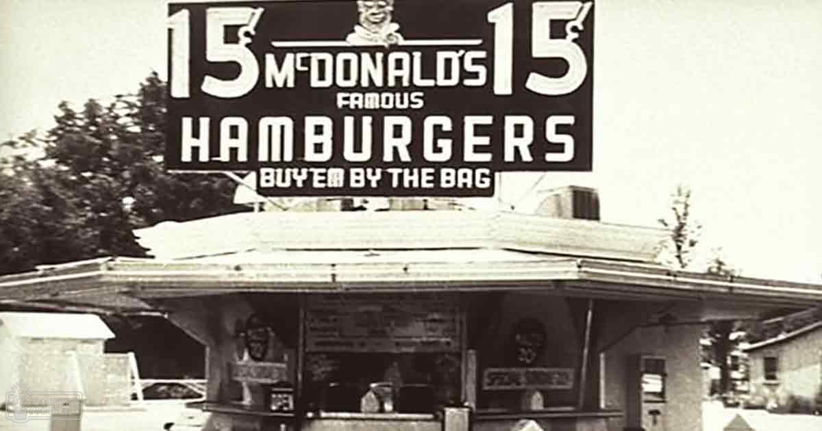 An old McDonald's restaurant from the mid-20th century advertising '15 Famous Hamburgers' for 15 cents each and 'Buy 'em by the Bag,' set outdoors with trees in spring or summer, under natural daylight.