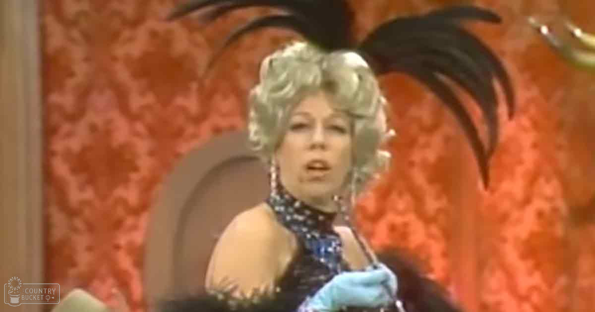 A middle-aged female performer in a theatrical setting, wearing an elegant black evening gown with sequin detailing from the 1950s to early 1960s. The scene includes ornate furniture and red patterned wallpaper.
