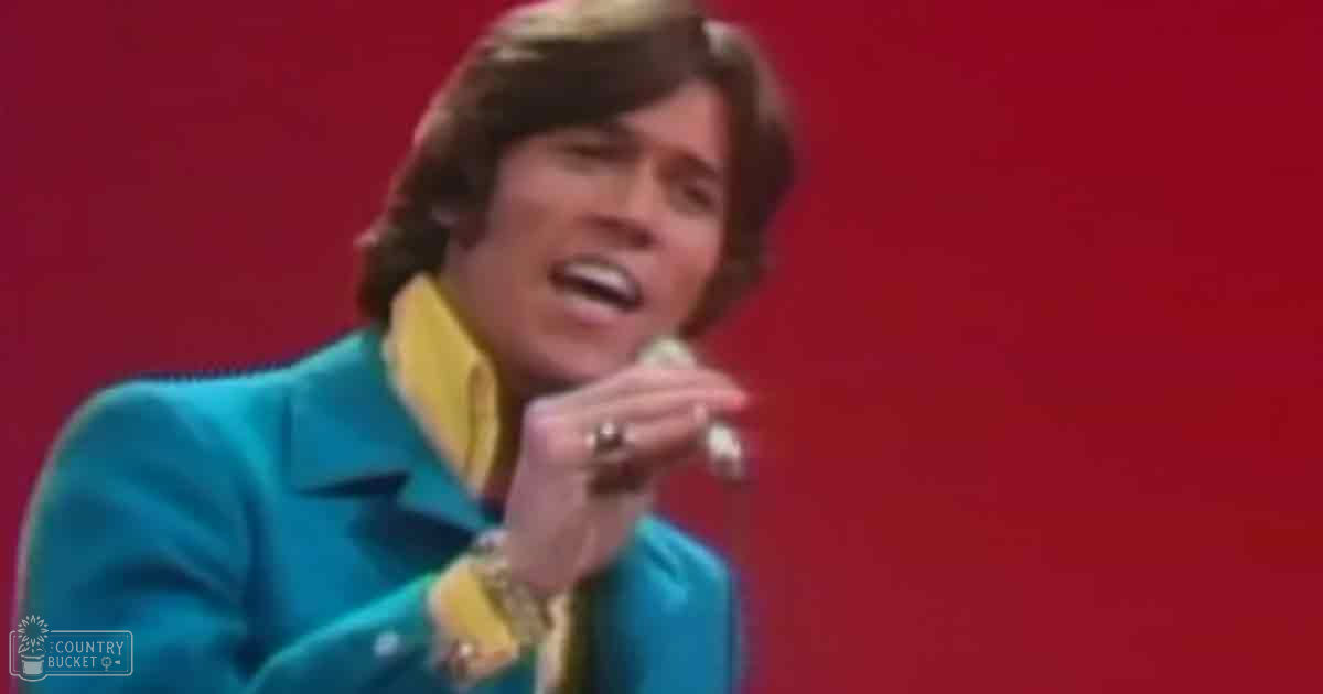 An adult male performer wearing a bright turquoise jacket with yellow shirt, holding a microphone against a red backdrop during an event or performance.