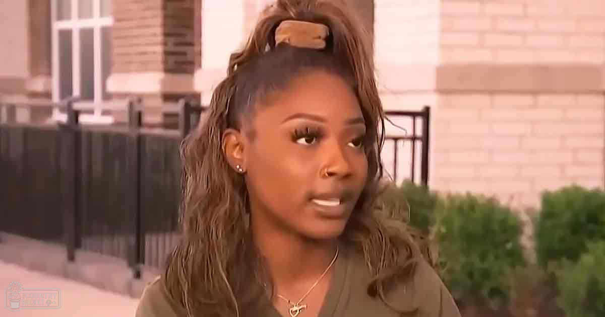 A young adult female wearing a relaxed olive green long-sleeved shirt with small stud earrings and a heart-shaped pendant necklace stands in an urban residential area, surrounded by brick buildings, windows, black metal fences, and green bushes under natural daylight.
