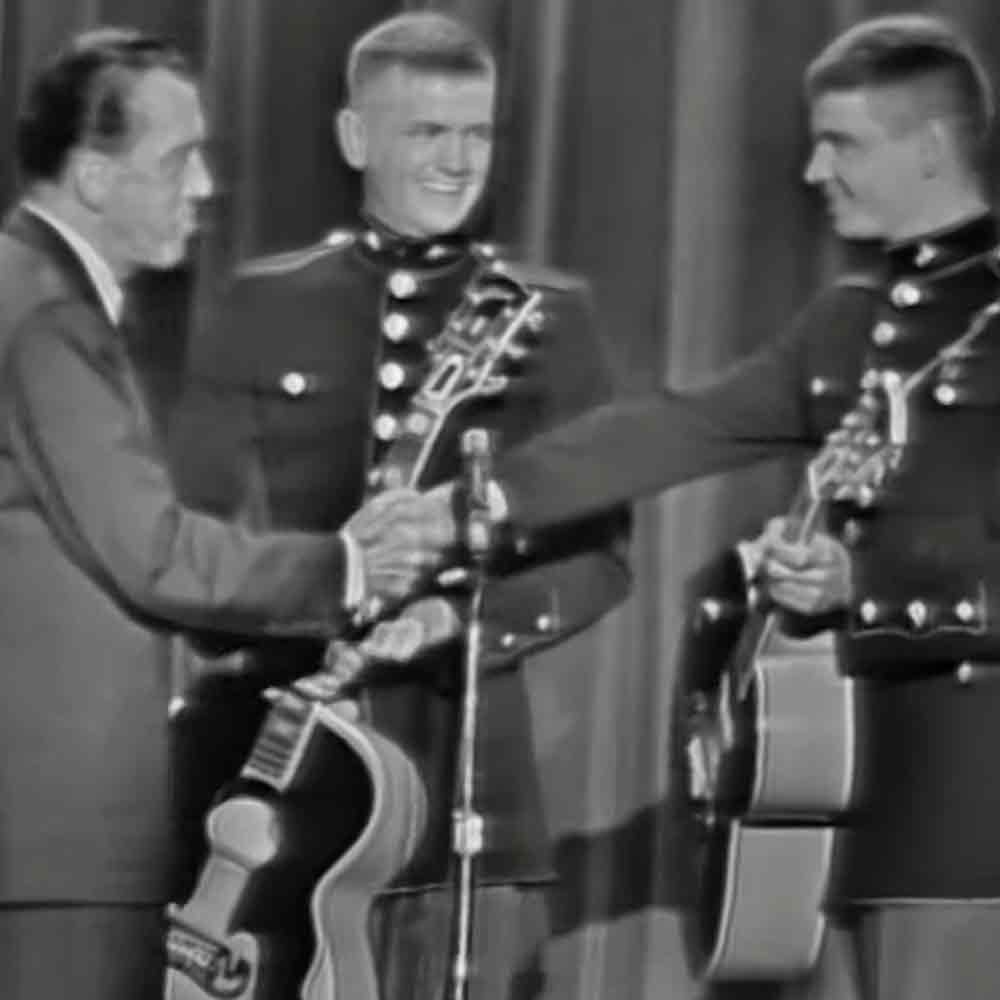 An adult male in a well-fitted suit jacket stands beside two young adults dressed in mid-20th century military uniforms, with musical instruments and stage equipment in the background.