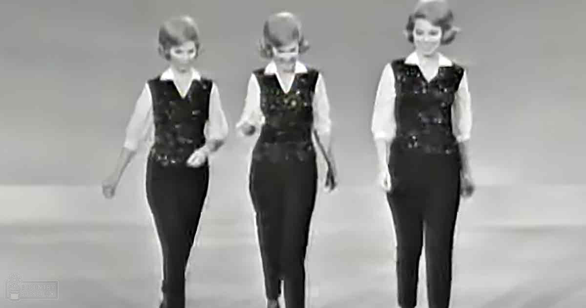 Group of young adult female dancers in a vintage dance routine, wearing white shirts and black patterned vests against a plain light-colored studio backdrop