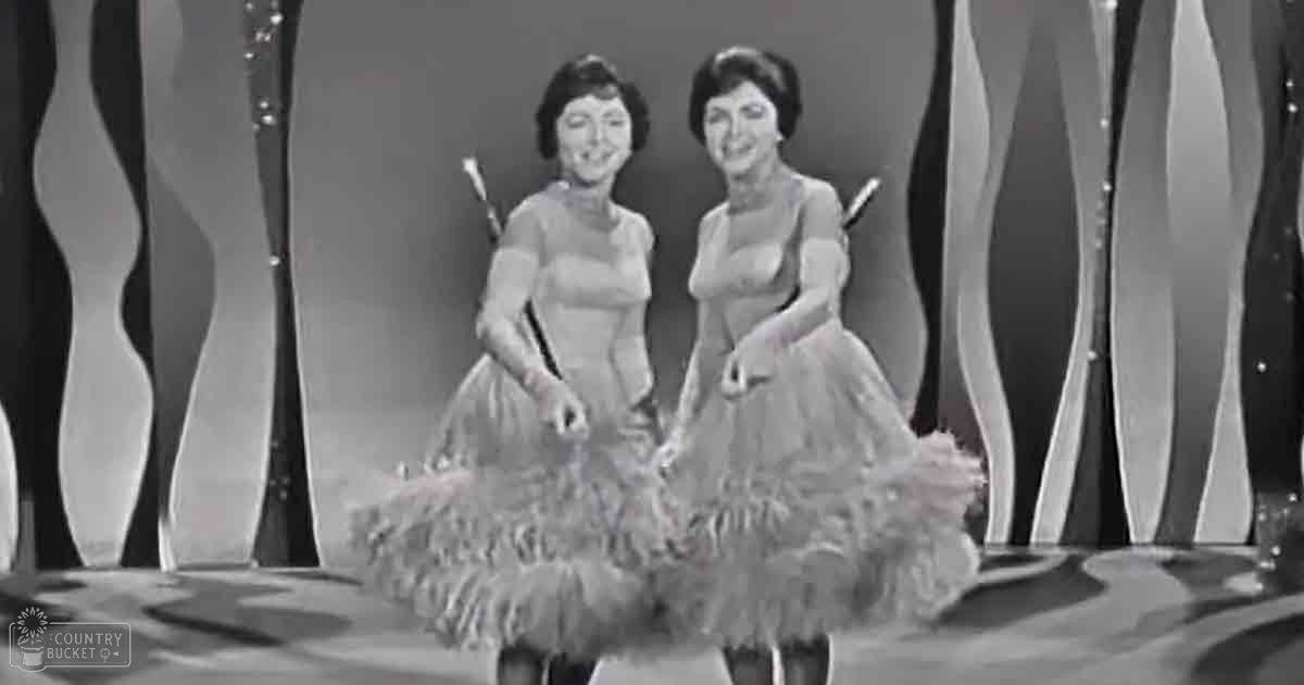 Two adult female performers wearing similar fringed dresses with short sleeves, holding wands or batons on a stage with abstract, wavy backdrop and decorative elements.