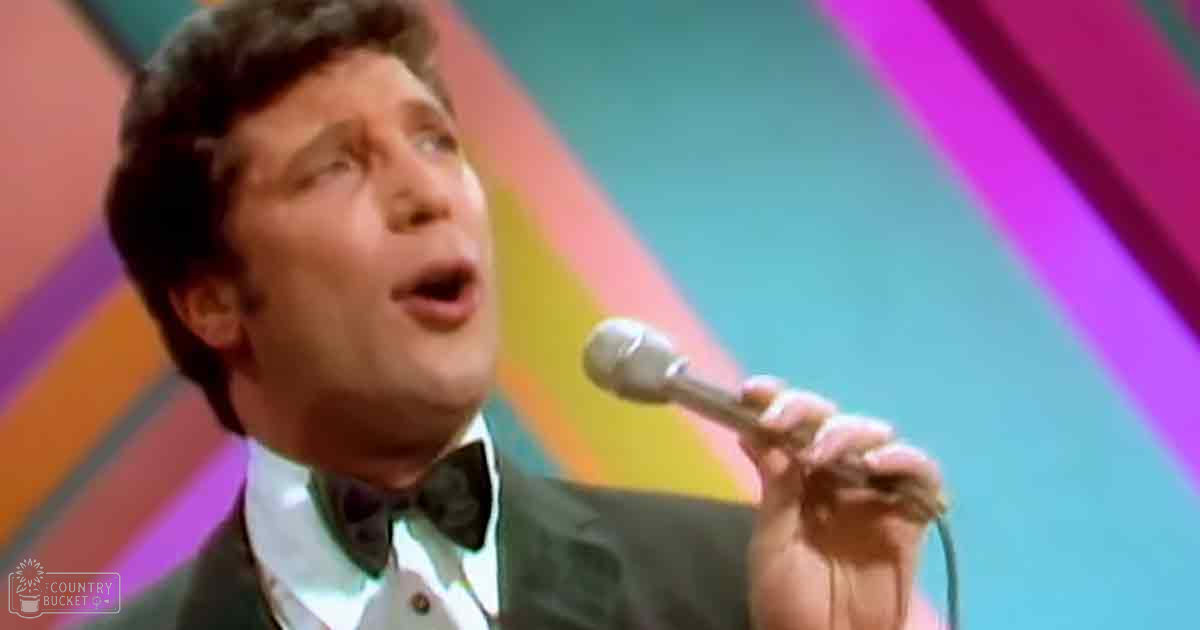 An adult male singer in a dark green suit jacket and white dress shirt with black bow tie performs on stage against a vibrant geometric backdrop.