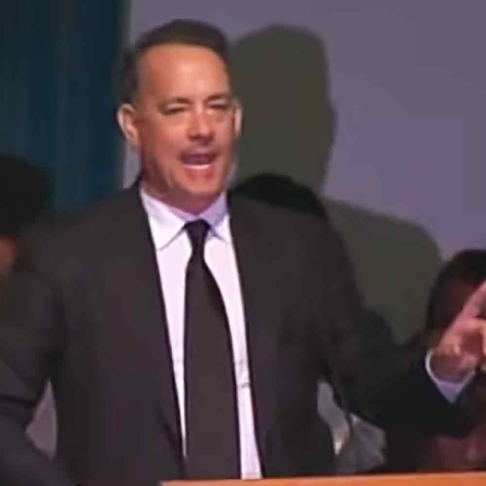 A middle-aged man at an event, dressed formally in a black suit jacket, white dress shirt with a black tie, surrounded by blurred figures of other people in dimly lit indoor space.