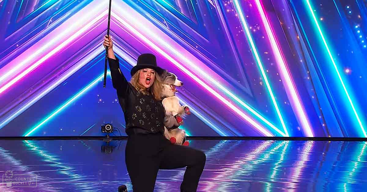 An adult female performer in a black outfit with small light-colored dots, holding a cane on an indoor stage illuminated by vibrant lighting.