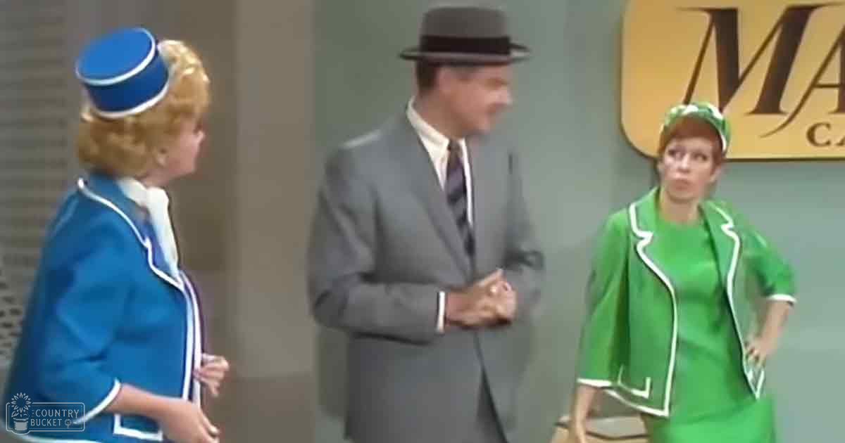 Adult male in a gray suit with a white shirt and dark tie, wearing a dark fedora hat. Young adult female flight attendant on the left in a blue uniform with white trim, and an adult female flight attendant on the right in a green uniform with white trim, standing indoors at what appears to be an airport.