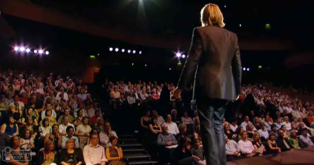 An adult male standing on stage addressing an audience in a theater or auditorium, surrounded by seated spectators dressed casually and formally for the occasion.